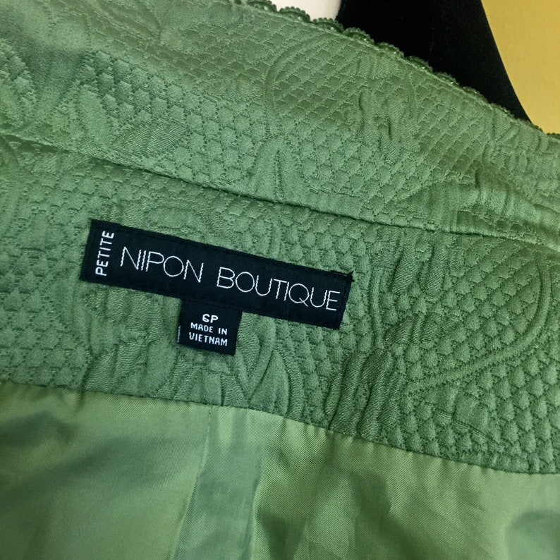 以下が含まれることがあります： 「PETITE NIPON BOUTIQUE」と「CP MADE IN VIETNAM」と書かれた黒いラベルが付いた緑色のキルティングジャケットです。