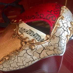 May include: A masquerade mask with a red, white and gold design. The mask has a cracked white finish with gold accents and musical notes. The mask has a black ribbon attached.