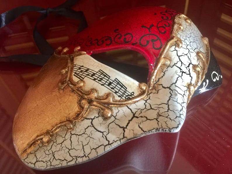 May include: A masquerade mask with a red, white and gold design. The mask has a cracked white finish with gold accents and musical notes. The mask has a black ribbon attached.
