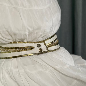 May include: A white dress with a gold and white chain belt. The belt has a buckle with two large metal studs.