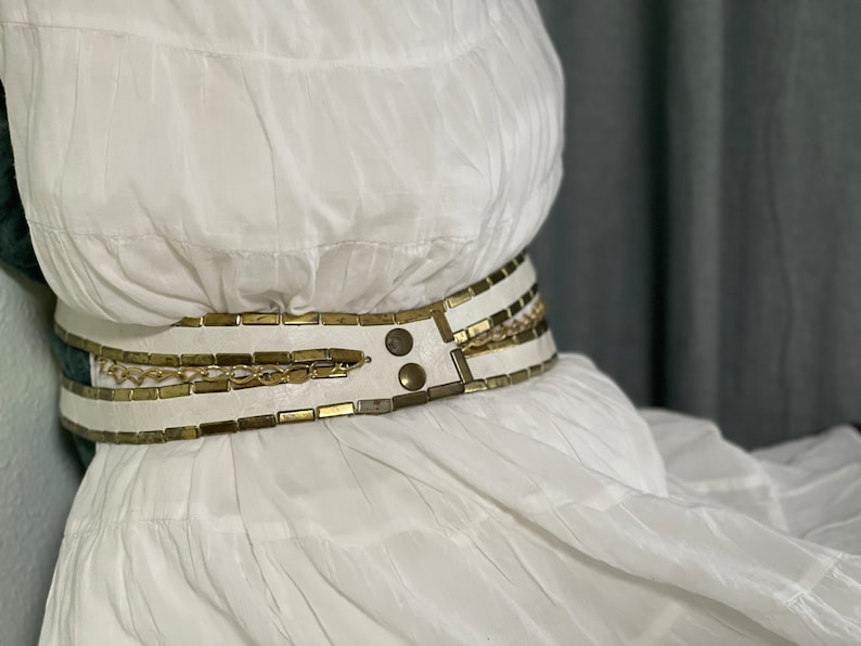 May include: A white dress with a gold and white chain belt. The belt has a buckle with two large metal studs.