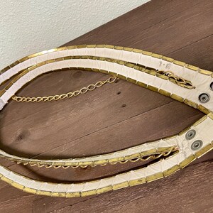 May include: A white leather belt with gold metal accents and a gold chain. The belt is made of individual squares of metal that are sewn together.