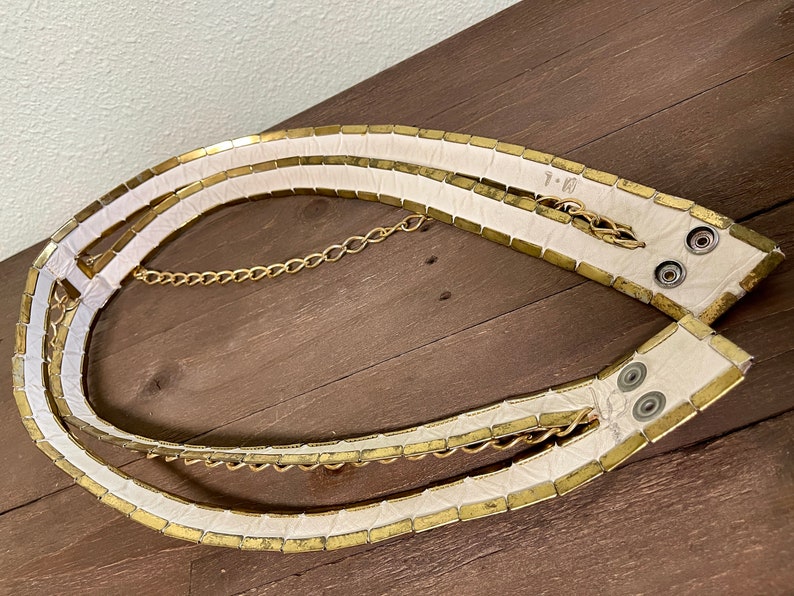May include: A white leather belt with gold metal accents and a gold chain. The belt is made of individual squares of metal that are sewn together.