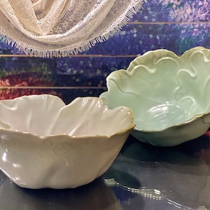 Puede incluir: Dos cuencos de cerámica con diseño de hoja. Uno es de color crema claro y el otro verde suave. Ambos cuencos tienen una superficie texturizada y un borde festoneado. Los cuencos se exhiben sobre una superficie oscura.