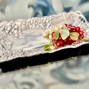 May include: A silver serving tray with a grapevine design. The tray is decorated with grapes and leaves. A bunch of red and white grapes is on the tray.