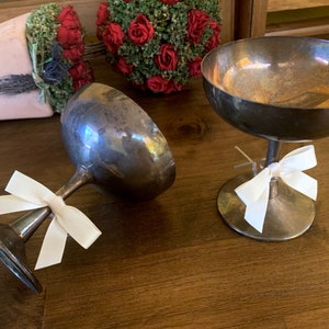 Puede incluir: Dos copas de champán plateadas con lazos de satén blanco. Las copas están sobre una superficie de madera.