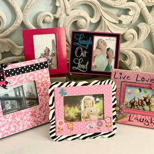 Può includere: Cinque cornici per foto rosa con design diversi. Le cornici sono decorate con pois, fiori, strisce zebrate e un fiocco. Le cornici sono tutte di dimensioni e forme diverse. Una cornice ha il testo "Laugh Out Loud". Un'altra cornice ha il testo "Live Love Laugh".