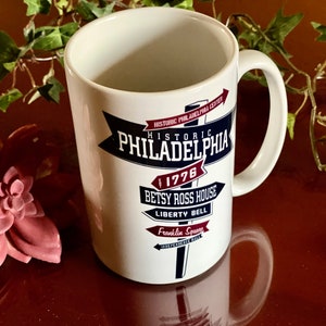 Puede incluir: Taza de cerámica blanca con un diseño azul y rojo que presenta el texto "Historic Philadelphia 1776" y una lista de monumentos históricos, incluyendo la casa de Betsy Ross, la Campana de la Libertad y Franklin Square.