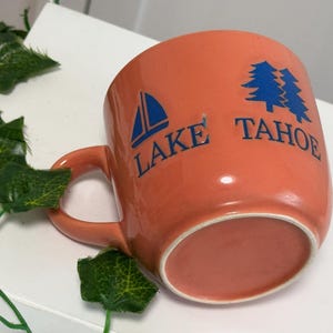 May include: An orange ceramic mug with the words "Lake Tahoe" in blue lettering. A blue sailboat and two blue pine trees are also printed on the mug. The mug has a curved handle.
