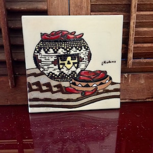 May include: A ceramic tile with a hand-painted design of a basket filled with red chili peppers. The basket is decorated with geometric patterns in brown, white, and yellow. The tile is signed "L. Kuhne" in the bottom right corner.