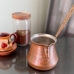 Puede incluir: Una cafetera de cobre con un acabado martillado y un mango decorativo. La cafetera está sobre una mesa junto a un frasco de vidrio con tapa de madera y una pequeña taza de café de cerámica.