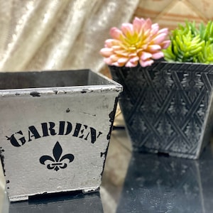 Könnte beinhalten: Zwei quadratische graue Pflanztöpfe. Der kleinere Topf ist weiß mit schwarzer Aufschrift "GARDEN" und einer schwarzen Fleur-de-lis. Der größere Topf ist schwarz mit einem Rautenmuster.