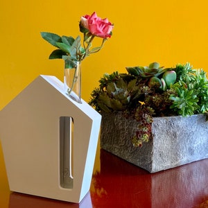 May include: A white house-shaped vase with a clear glass tube insert, holding a single pink rose. Next to the vase is a gray planter filled with green succulents.