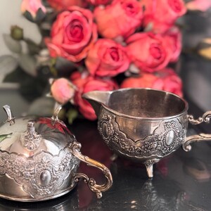 K&ouml;nnte beinhalten: Ein silbernes Sahnegie&szlig;er- und Zuckergef&auml;&szlig;-Set mit aufwendigen Blumendesigns. Der Sahnegie&szlig;er hat einen Griff und einen Ausguss, w&auml;hrend das Zuckergef&auml;&szlig; einen Deckel hat.