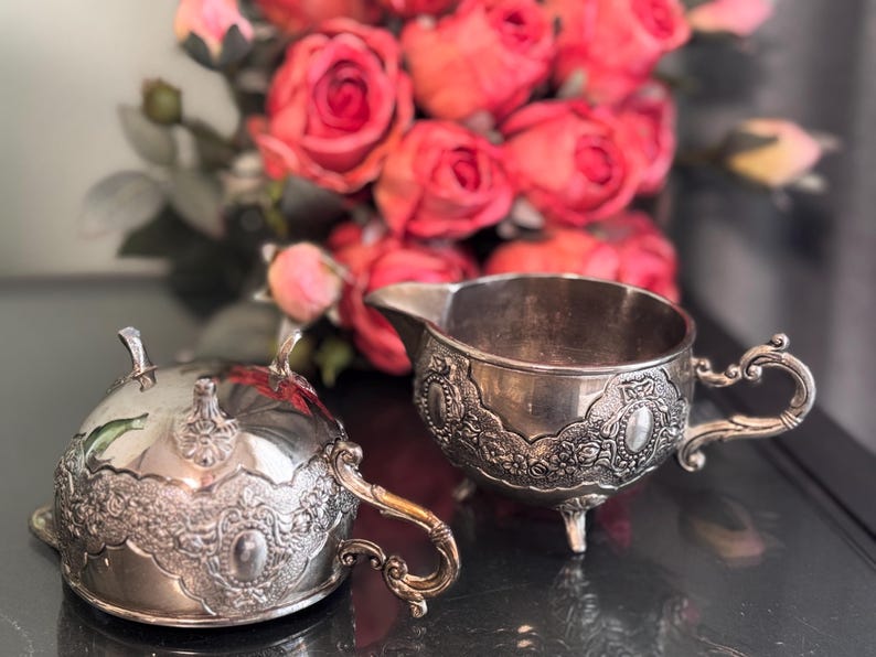 K&ouml;nnte beinhalten: Ein silbernes Sahnegie&szlig;er- und Zuckergef&auml;&szlig;-Set mit aufwendigen Blumendesigns. Der Sahnegie&szlig;er hat einen Griff und einen Ausguss, w&auml;hrend das Zuckergef&auml;&szlig; einen Deckel hat.