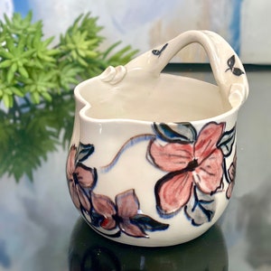 May include: A white ceramic bowl with a floral design. The bowl has a handle and is decorated with pink, blue, and green flowers.