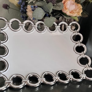 May include: A rectangular white serving tray with a decorative silver chain-link border. The tray is made of a glossy material and is suitable for serving food or displaying decorative items. The tray is a stylish addition to any home decor.