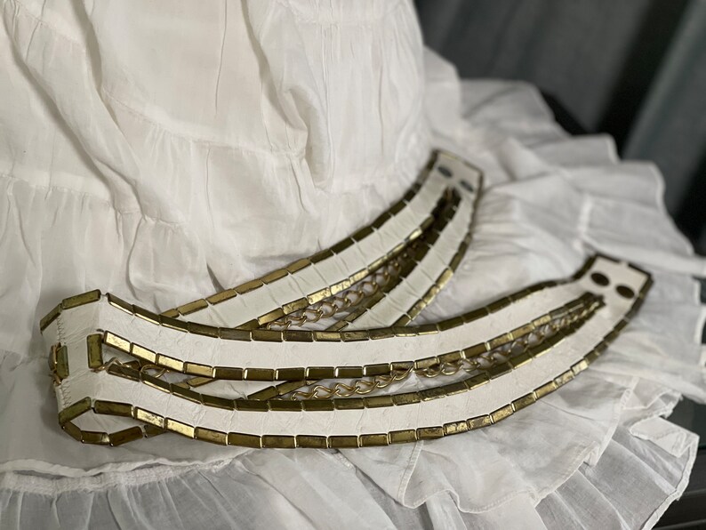 May include: A white leather belt with gold chain accents. The belt is made of multiple strips of leather with gold metal accents.