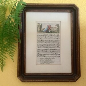 May include: A framed vintage print of a musical score with a small illustration of a couple in a garden. The text reads "C'est l'amour qui nous rend heureux" and includes musical notes.