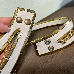 May include: White leather belt with gold-toned metal accents and a chain detail. The belt is made of rectangular pieces of metal with a white leather backing.
