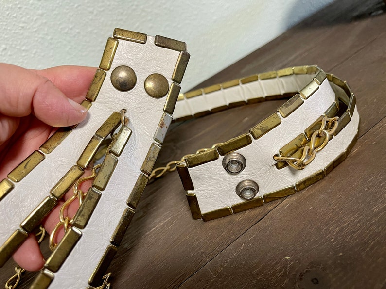 May include: White leather belt with gold-toned metal accents and a chain detail. The belt is made of rectangular pieces of metal with a white leather backing.