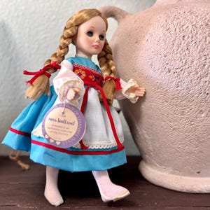 May include: A vintage doll, "Miss Holland," dressed in a traditional Dutch costume. The doll has blonde braids tied with red ribbons, a blue skirt, and a white blouse with red and floral accents. A tag reads "World of Celebrated Dolls."