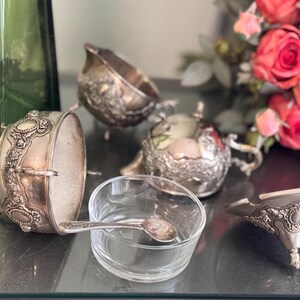 K&ouml;nnte beinhalten: Ein Satz silberplattierter Servierplatten mit aufwendigen Blumendesigns. Die Platten stehen auf einem Tisch mit einer Glassch&uuml;ssel und einem L&ouml;ffel.