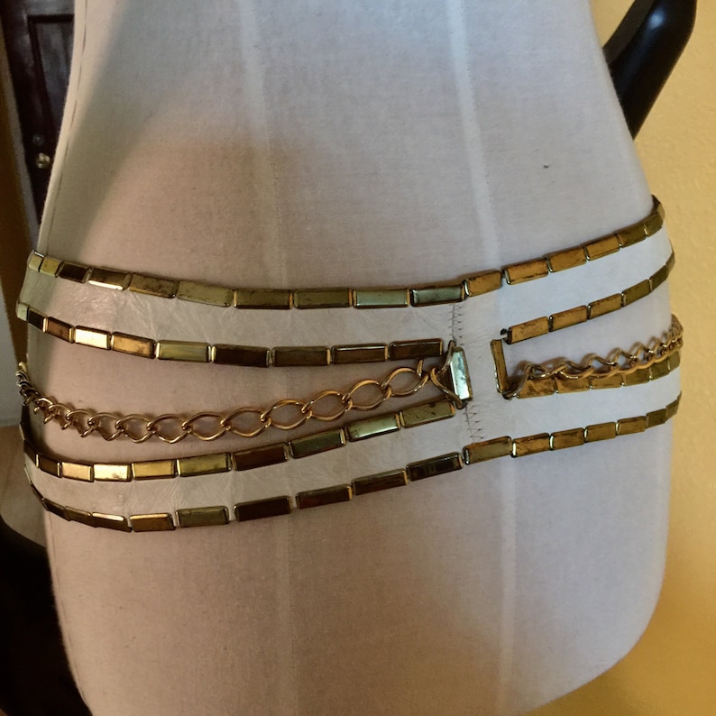May include: A white leather belt with gold-toned metal rectangular links and a chain. The belt is layered with three rows of rectangular links and one row of chain.