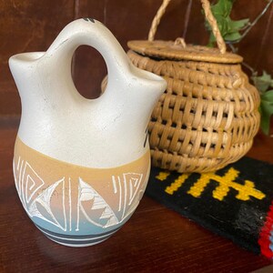 May include: A white and tan ceramic vase with a distinctive handle, decorated with geometric patterns in white, tan, and blue. A small woven basket and a black and yellow woven mat are in the background.