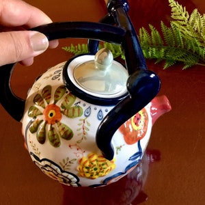Puede incluir: Una tetera de cerámica blanca con asa y tapa azules. La tetera está decorada con flores y hojas de colores.