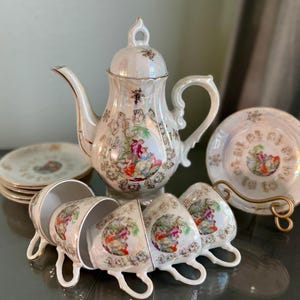 Könnte beinhalten: Ein Vintage-Porzellan-Tee-Set mit floralem Design. Das Set enthält eine Teekanne, vier Tassen und vier Untertassen. Die Teekanne hat einen Deckel mit einem Knopf in Form einer Blume. Die Tassen und Untertassen haben ein zartes Blumenmuster mit goldenen Akzenten.