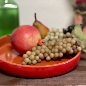 Puede incluir: Un cuenco de cerámica rojo lleno de una variedad de frutas, incluyendo una granada, una pera y uvas verdes.