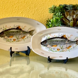 Puede incluir: Dos platos ovalados blancos con ribete dorado, cada uno con una imagen pintada a mano de un pez con detalles verdes, marrones y amarillos. Los platos están sobre soportes negros.