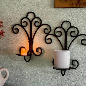 May include: Two black wrought iron wall sconces with a scrolled design. One holds a lit candle, the other a white cylindrical container. The sconces are mounted on a textured, light green wall, creating a warm, inviting ambiance.