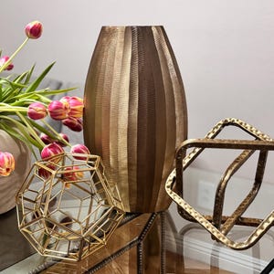 May include: A decorative arrangement featuring a tall, textured bronze vase, geometric gold sculptures, and a vase of pink and yellow tulips. The objects are arranged on a glass surface.