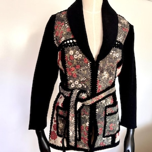 May include: A black knit cardigan with a floral patchwork design. The cardigan has a wide collar, a belt, and two pockets.