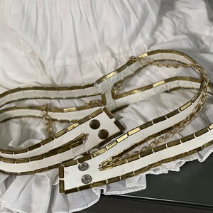 May include: A white leather belt with gold metal accents. The belt is made of multiple layers of leather and has a chain detail.