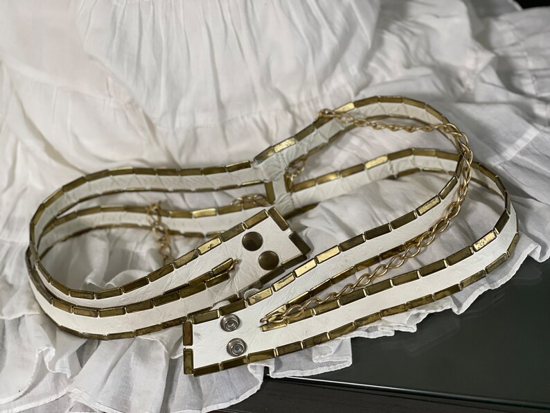May include: A white leather belt with gold metal accents. The belt is made of multiple layers of leather and has a chain detail.