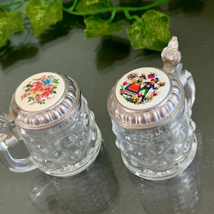 以下が含まれることがあります： 銀の蓋が付いた小さなガラス製のビアジョッキが2つ。1つのジョッキは蓋に花柄のデザインが施され、もう1つは伝統的なドイツの衣装を着たカップルが描かれています。