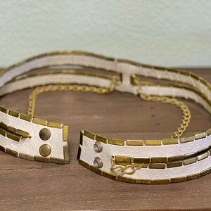 May include: A white leather belt with gold metal accents and a chain. The belt is made of multiple layers of leather and has a chain that runs through the center.