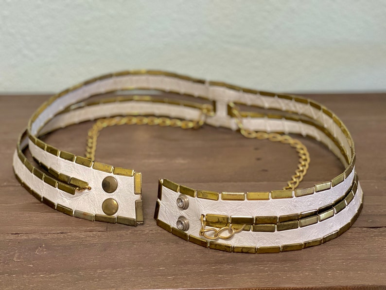 May include: A white leather belt with gold metal accents and a chain. The belt is made of multiple layers of leather and has a chain that runs through the center.