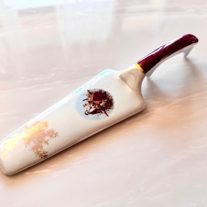 May include: A white porcelain cake server with a burgundy handle and gold accents. The server features a floral design and a small image of a couple in the center.