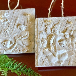 May include: Two white ceramic wall plaques with a raised design of fairies in a garden setting. Each plaque has a string for hanging.