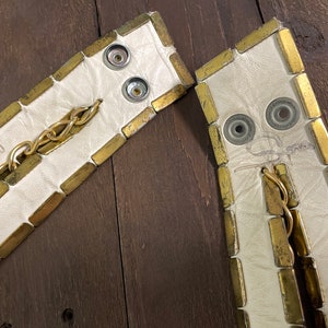 May include: Two white leather straps with gold metal accents. The straps have a chain and metal snaps. The straps are marked with the letters "T.M."