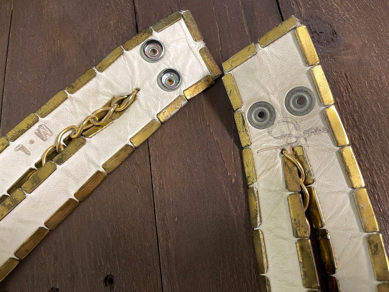 May include: Two white leather straps with gold metal accents. The straps have a chain and metal snaps. The straps are marked with the letters "T.M."