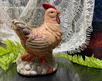 Pode incluir: Estatueta de galo de cerâmica em tons de bege, castanho e vermelho. O galo tem uma crista e barbela vermelhas, com um design detalhado de penas. A base tem uma aparência texturizada, semelhante à pedra. Um item decorativo para uma cozinha ou decoração de quinta.