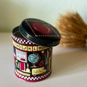 Può includere: Una latta decorativa con coperchio con un design a mela rossa. La latta cilindrica è decorata con grafiche a tema scolastico, tra cui un mappamondo, libri e un autobus scolastico. Il coperchio è leggermente socchiuso, rivelando l'interno.