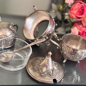 K&ouml;nnte beinhalten: Ein silbernes Zucker- und Milchk&auml;nnchen-Set mit aufwendigen Blumendesigns. Das Set enth&auml;lt eine Zuckerdose mit Deckel, einen Milchk&auml;nnchen und einen kleinen L&ouml;ffel. Alle Teile sind aus Silber gefertigt und haben eine polierte Oberfl&auml;che.