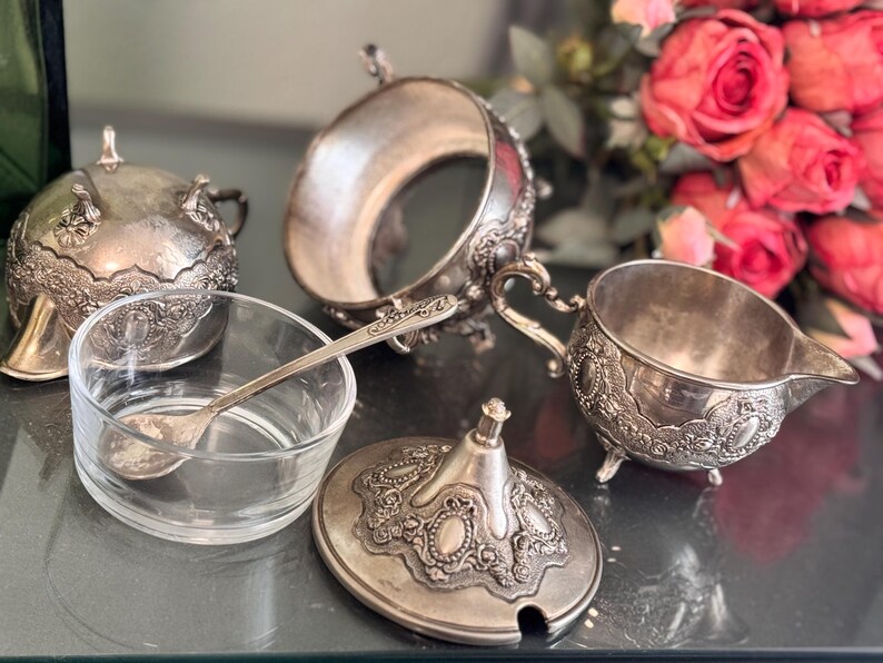 K&ouml;nnte beinhalten: Ein silbernes Zucker- und Milchk&auml;nnchen-Set mit aufwendigen Blumendesigns. Das Set enth&auml;lt eine Zuckerdose mit Deckel, einen Milchk&auml;nnchen und einen kleinen L&ouml;ffel. Alle Teile sind aus Silber gefertigt und haben eine polierte Oberfl&auml;che.