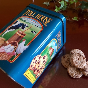 May include: A teal Toll House cookie tin with a farm scene illustration and the word "Nestle". Several chocolate chip cookies are next to the tin. The tin is a limited edition.